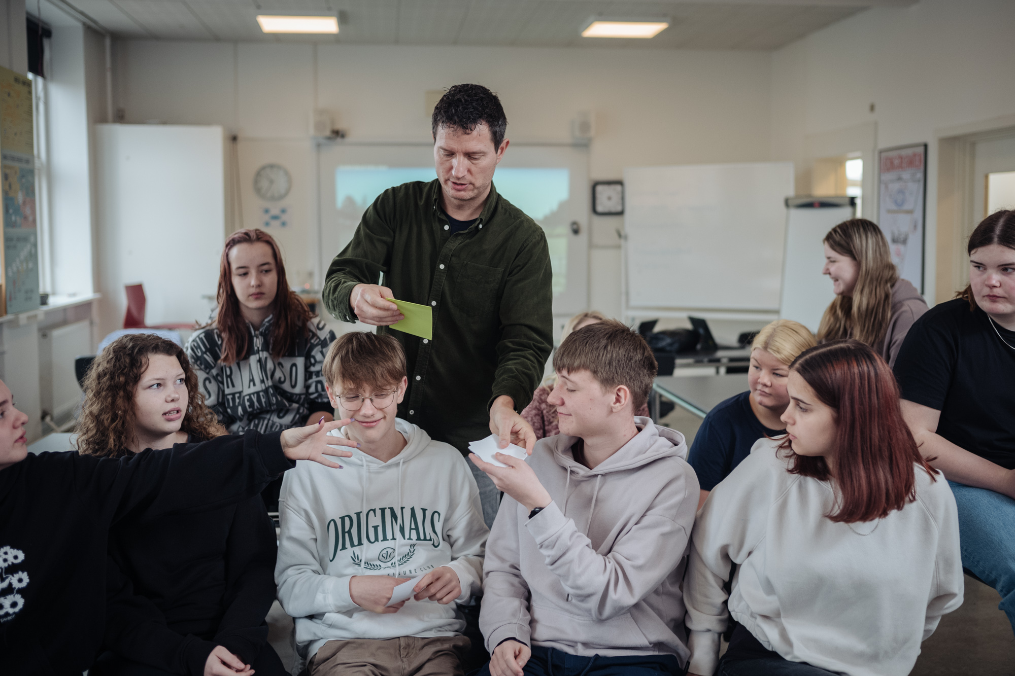 Billede af undervisningsituation med skolelederen og en gruppe elever, der modtager papirlapper.